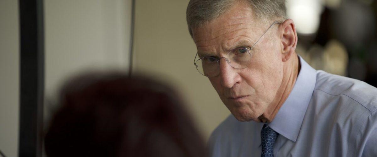 En ældre mand med kort, gråt hår, briller og blå skjorte med slips kigger intenst på en person, der står med ryggen til kameraet. Omgivelserne ser ud til at være et indendørs kontor eller mødelokale.