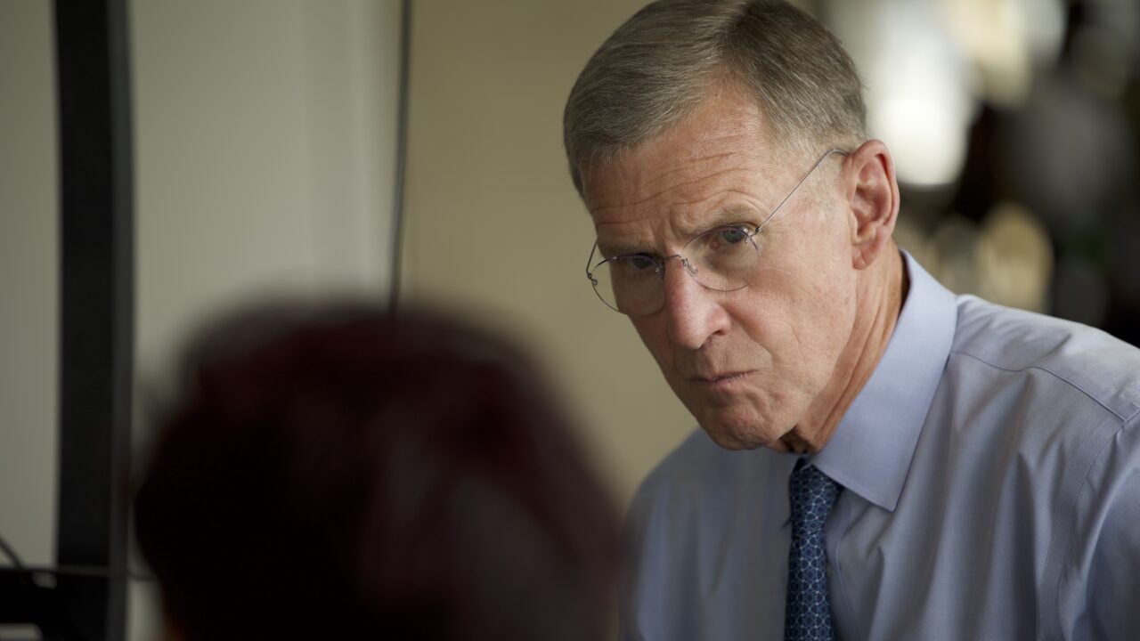 En ældre mand med kort, gråt hår, briller og blå skjorte med slips kigger intenst på en person, der står med ryggen til kameraet. Omgivelserne ser ud til at være et indendørs kontor eller mødelokale.