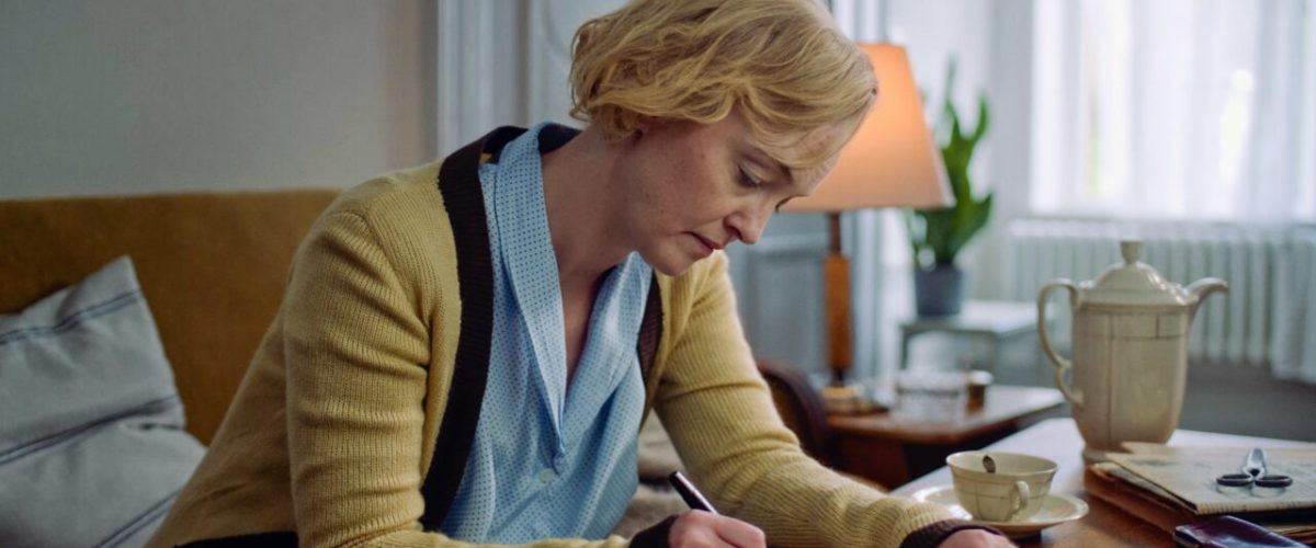 En kvinde med kort blondt hår, iført blå top og beige cardigan, sidder ved et bord og skriver i en notesbog. En tekande, en kop og en lampe står på bordet i et veloplyst rum med store vinduer.