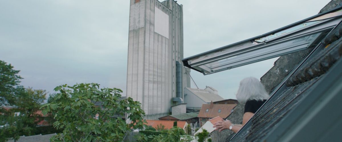 En person med gråt hår læner sig ud af et tagvindue og kigger mod en høj, grå industribygning med små vinduer. Træer og beboelseshuse er synlige nedenfor under en overskyet himmel.