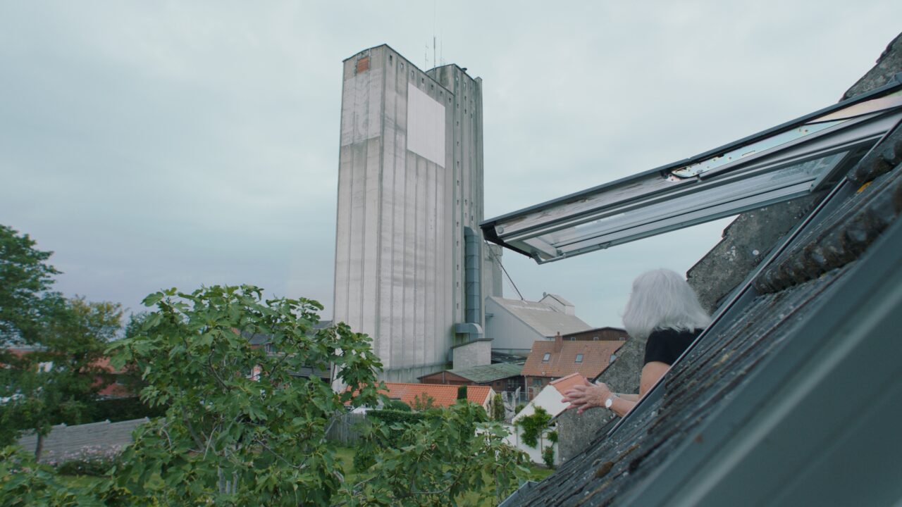 En person med gråt hår læner sig ud af et tagvindue og kigger mod en høj, grå industribygning med små vinduer. Træer og beboelseshuse er synlige nedenfor under en overskyet himmel.