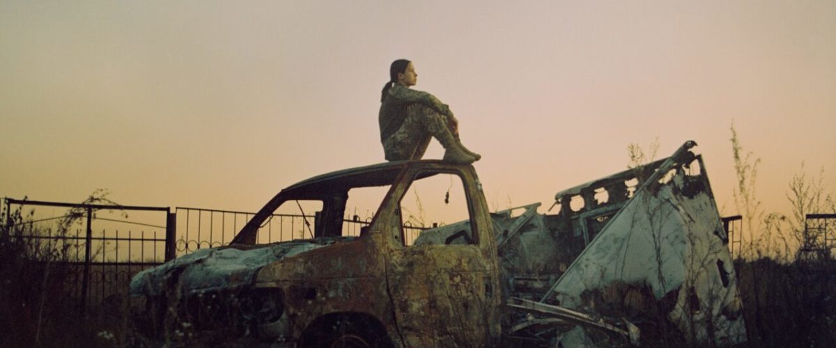 En person sidder på taget af en rusten, udbrændt bil på en tilgroet mark nær et metalhegn i skumringen. Himlen er en svag lysgradient, der antyder tidlig aften eller daggry.