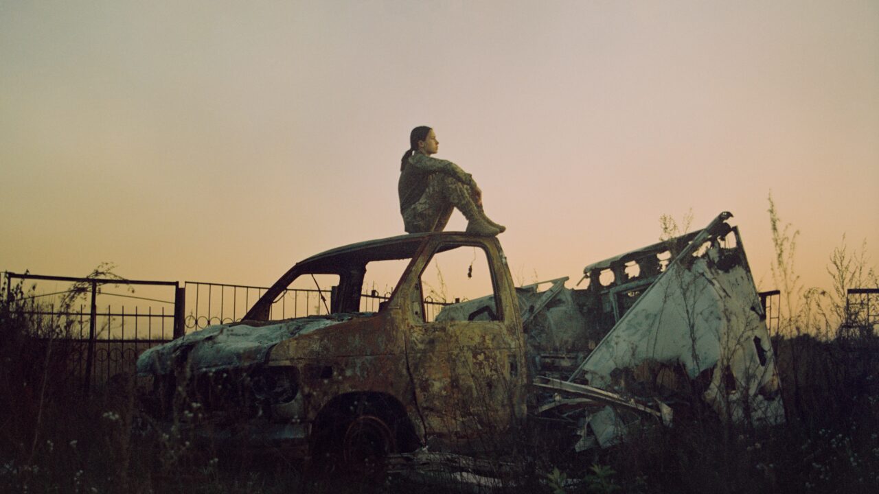 En person sidder på taget af en rusten, udbrændt bil på en tilgroet mark nær et metalhegn i skumringen. Himlen er en svag lysgradient, der antyder tidlig aften eller daggry.