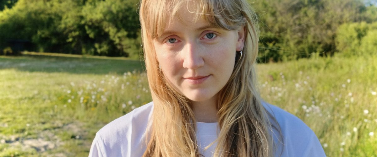 En ung kvinde med langt lyst hår og en hvid skjorte står udendørs i et solbeskinnet græsareal med træer og buske i baggrunden. Hun ser direkte ind i kameraet med et neutralt udtryk.