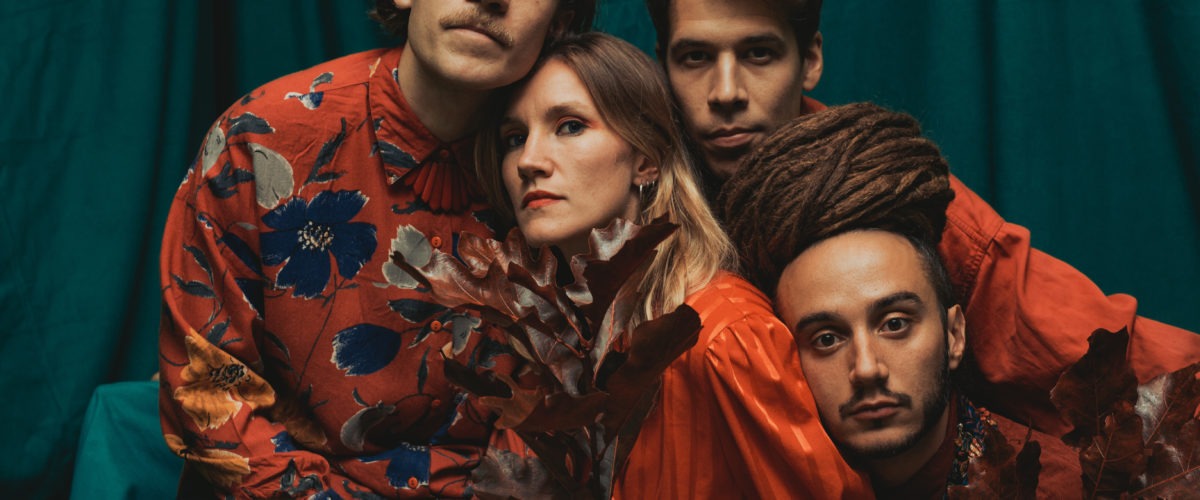 Fire personer poserer tæt sammen foran en blågrøn stofbaggrund, alle iført rødmønstrede skjorter. To holder tørre, brune blade. En person har lange dreadlocks, og en anden har overskæg. De ser direkte ind i kameraet.