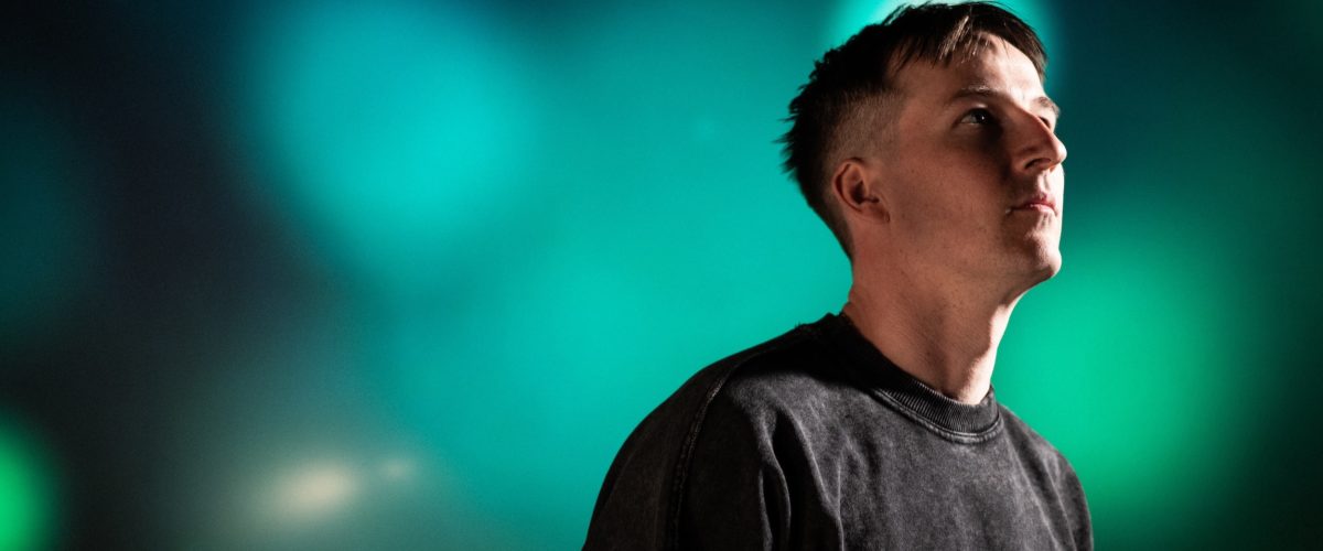 En person med kort hår iført en mørk sweatshirt kigger opad med en sløret baggrund af grønne og blå lys.