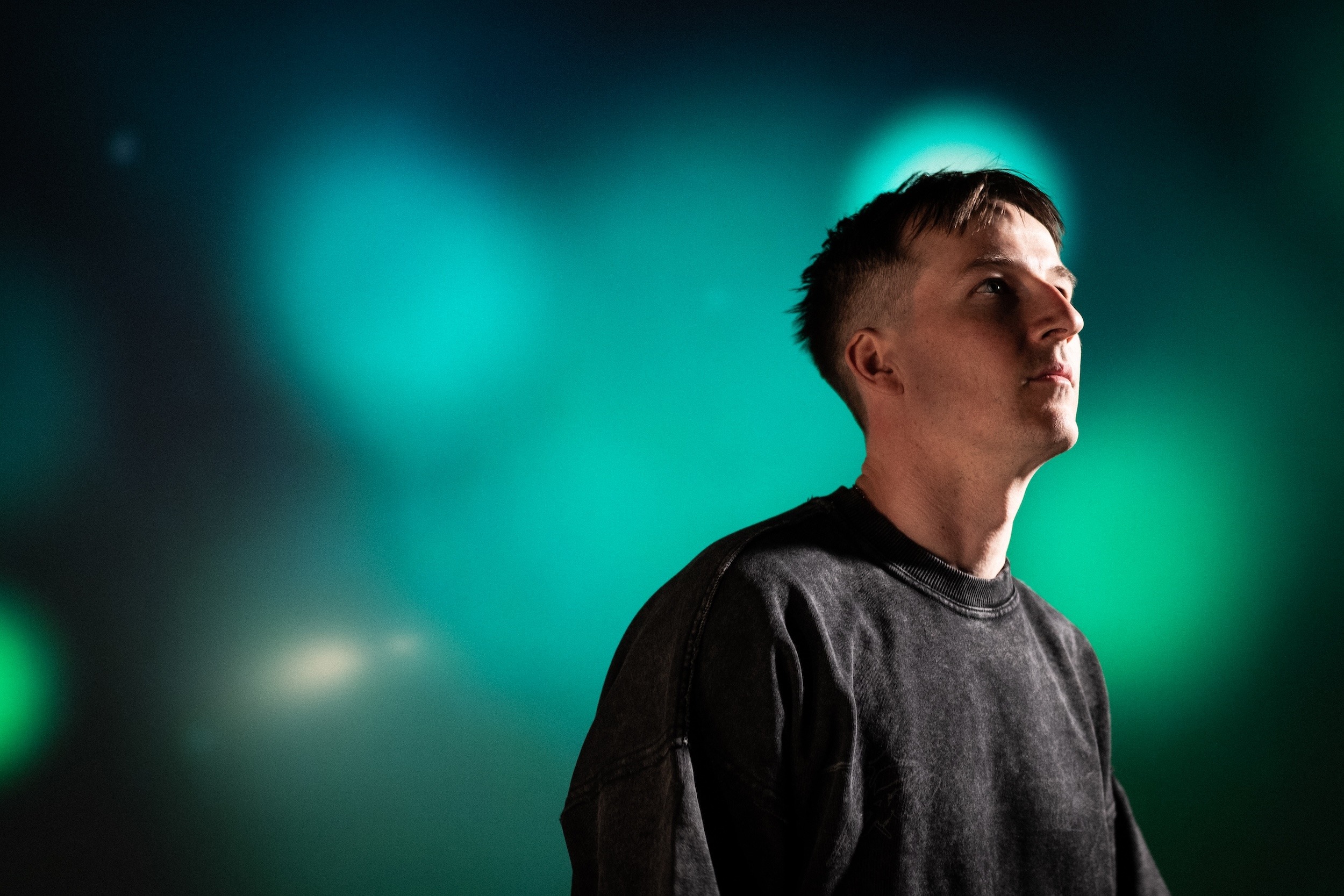 En person med kort hår iført en mørk sweatshirt kigger opad med en sløret baggrund af grønne og blå lys.