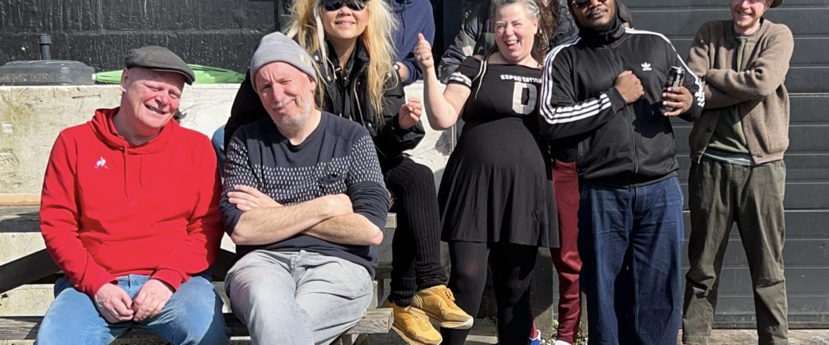 En gruppe på otte voksne poserer udendørs på trætrin og på jorden, mens nogle smiler og andre laver legende bevægelser. De har afslappet tøj og hatte på. I baggrunden ses en sort bygning og et indhegnet område.