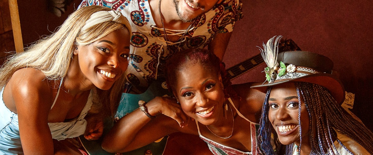 Fire personer smiler op til kameraet, mens de er samlet omkring et keyboard, en violin og trommestikker. De ser ud til at være i et varmt oplyst rum, muligvis et musikstudie. En person bærer en hat med dekorative fjer.