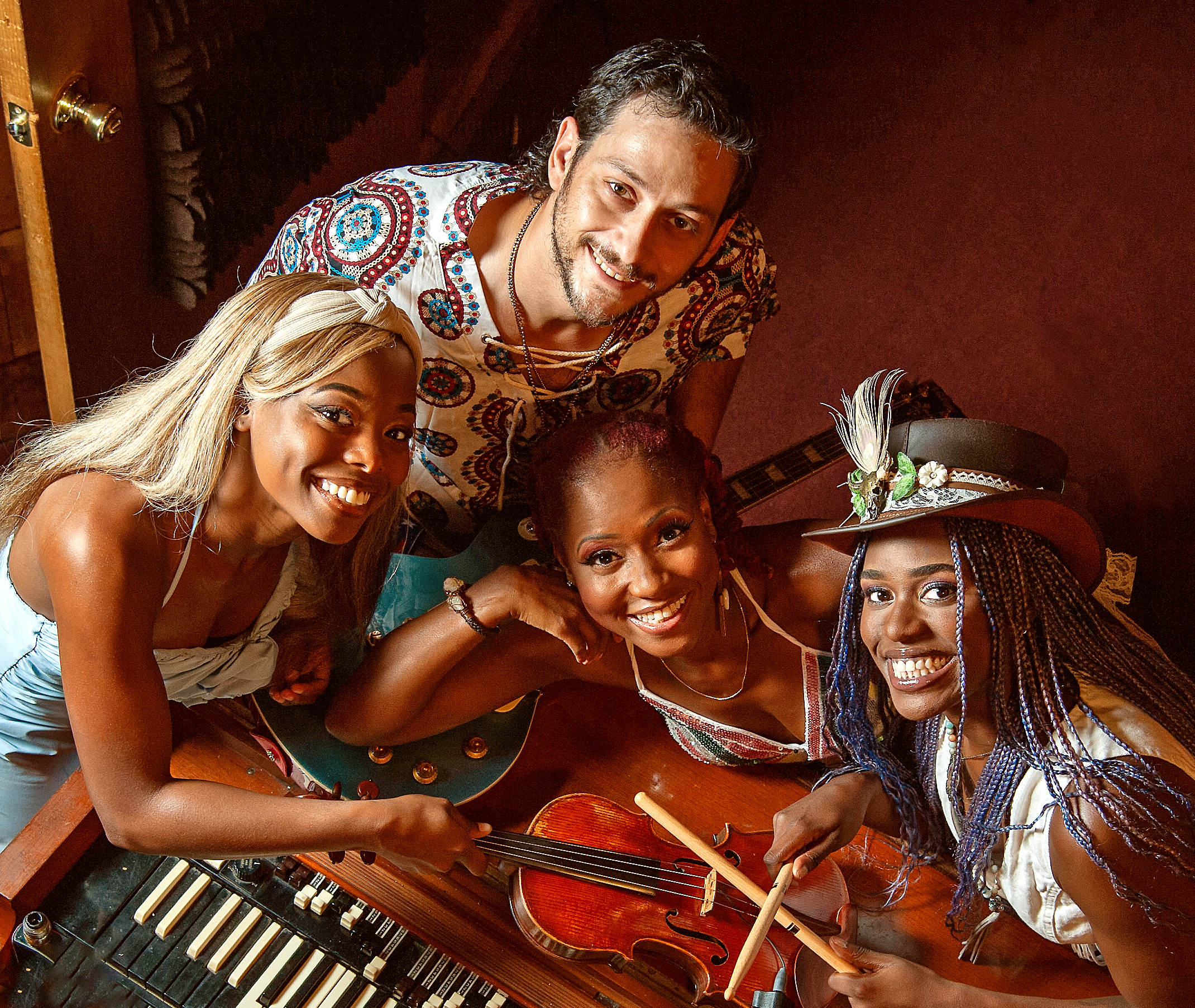 Fire personer smiler op til kameraet, mens de er samlet omkring et keyboard, en violin og trommestikker. De ser ud til at være i et varmt oplyst rum, muligvis et musikstudie. En person bærer en hat med dekorative fjer.