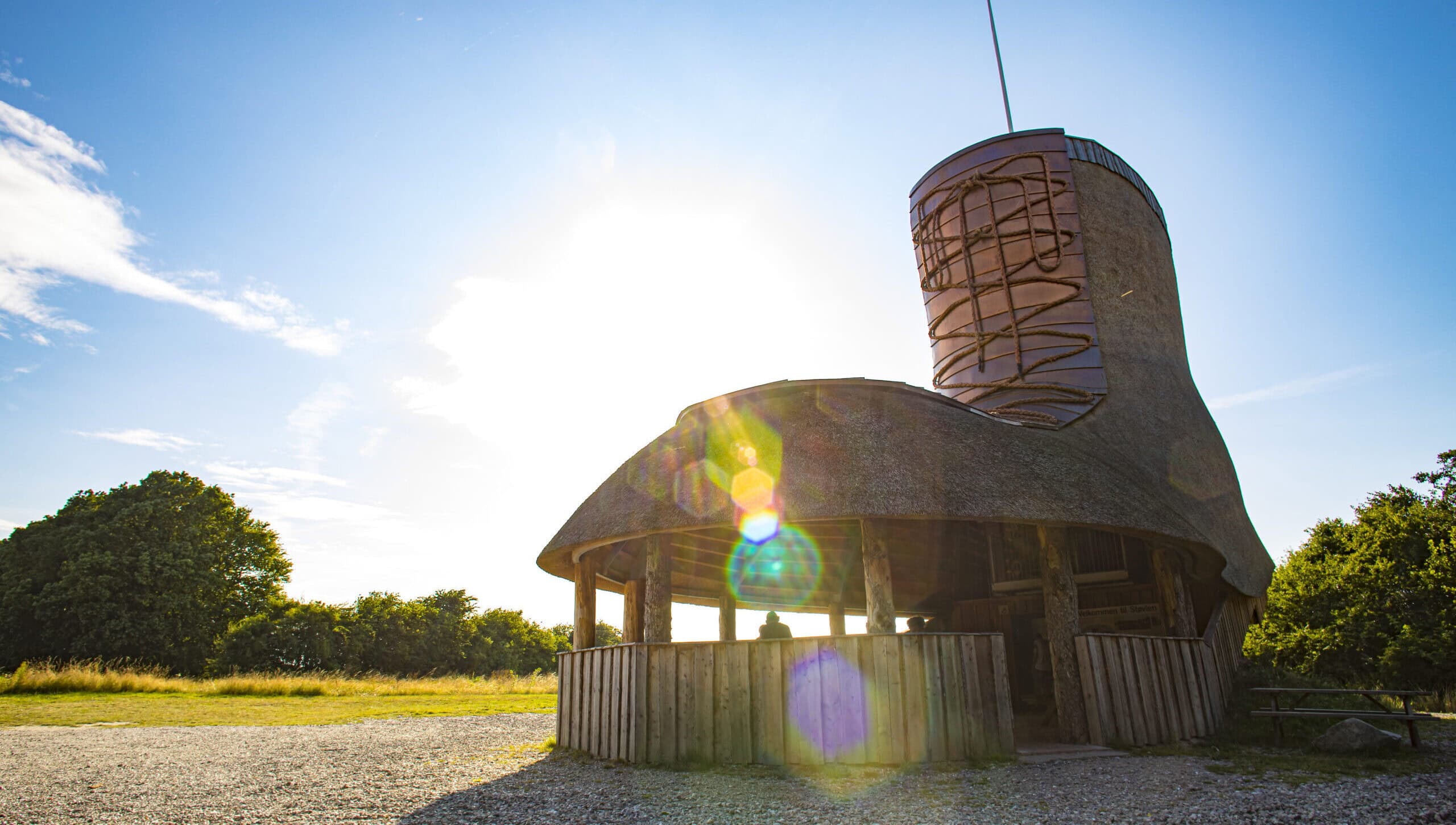 En stor, finurlig træstruktur formet som en støvle står under en lyseblå himmel med sollys og refleksioner. Den er omgivet af træer og græsklædte marker med et grusområde i forgrunden. En stor, finurlig træstruktur formet som en støvle står under en lyseblå himmel med sollys og refleksioner. Den er omgivet af træer og græsklædte marker med et grusområde i forgrunden.