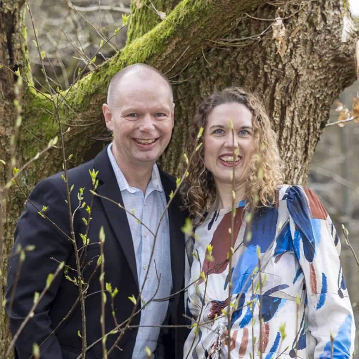 To personer foran et træ, smilende, i naturen, til en kulturel begivenhed.